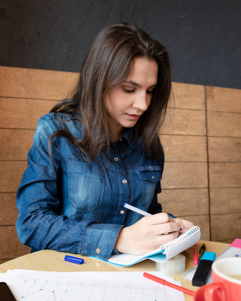 Girl in a blue denim shirt sitting in a cafe makes notes in a notepad with a pen. A smartphone, sheets with various charts, a red mug with green tea, felt-tip pens are on the table - Foto, Imagen