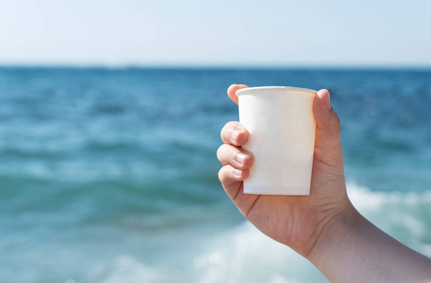 若い女性の手は青い海の背景に白い紙コップを持っています明るい夏の日にテキストのためのコピースペース。アクティブなライフコンセプト、旅行や休暇. - 写真・画像