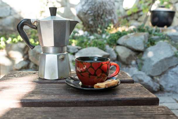 Чашка кави і залізна гейзерна кавоварка стоїть на темному старому дерев'яному столі зовні в яскравий літній день. Ранкова релаксація, активний спосіб життя, насолоджуючись кемпінгом
 - Фото, зображення