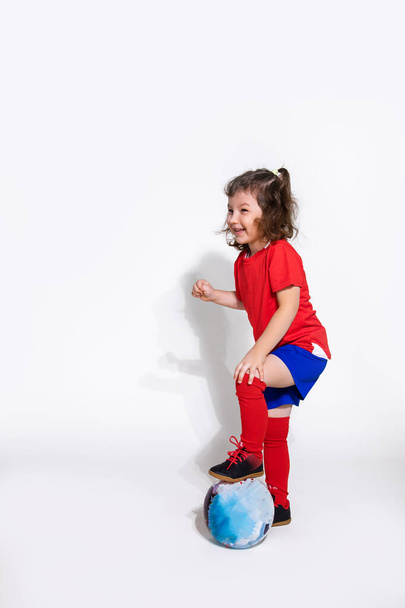 Little pretty girl in a football uniform and with ball on awhite background looks to side. Women's football concept for kids - Fotografie, Obrázek