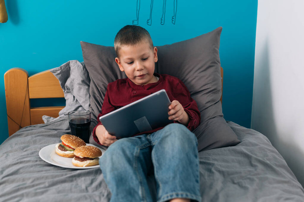 niño comiendo alimentos poco saludables y el uso de tabletas digitales mientras está sentado en su habitación - Foto, imagen