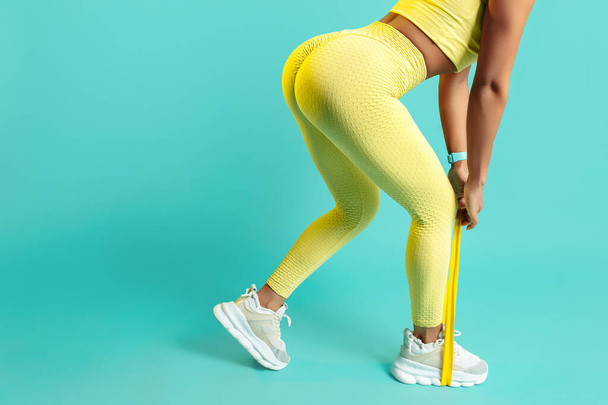 Close-up photo of sporty young woman in yellow sport outfit doing exercises with stretch band over blue background. - Φωτογραφία, εικόνα