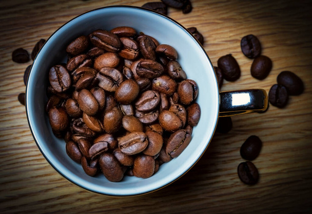 Φρέσκοι κόκκοι καφέ σε ένα φλιτζάνι espresso με ασημένια λαβή σε ξύλινο τραπέζι περιτριγυρισμένο από διάσπαρτους κόκκους καφέ - Φωτογραφία, εικόνα