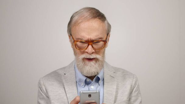 Homem idoso irritado usando telefone no estúdio. Focado cara colocando em óculos dentro de casa. - Foto, Imagem