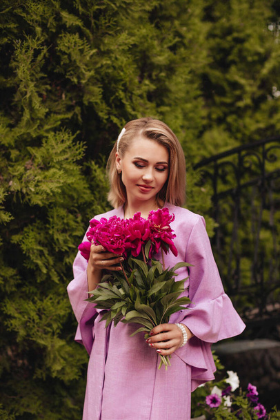 Eine schöne junge Frau in einem rosafarbenen Leinenkleid hält rosa Pfingstrosen in den Händen. Mädchen mit einem Blumenstrauß. Lifestyle-Konzept.  - Foto, Bild
