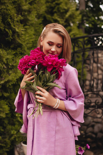 A beautiful young woman in a pink linen dress holds pink peonies in hands. Girl with a bouquet of flowers. Lifestyle concept.  - Fotografie, Obrázek