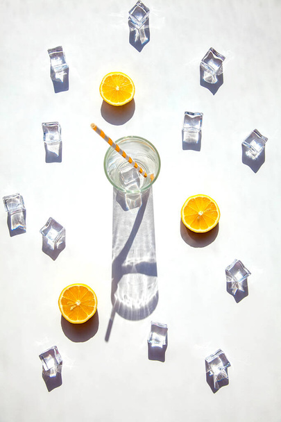 Ένα άδειο ποτήρι με μια μεγάλη σκιά γύρω του, παγάκια και φέτες πορτοκαλιού σε λευκό φόντο. Ηλιακή φωτογραφία. Top view, ζεστό καλοκαίρι και ποτό έννοια. - Φωτογραφία, εικόνα
