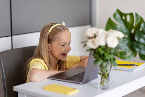 Cute little caucasian girl with blonde hair in fashionable dress illuminating yellow color sitting at home during coronavirus pandemic quarantine and using laptop and mobile phone. Stay at home during - Photo, image