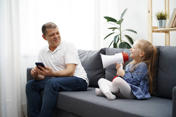Grandpa sitting on sofa and using smartphone, little granddaughter shouting through megaphone to him to stop - Foto, afbeelding