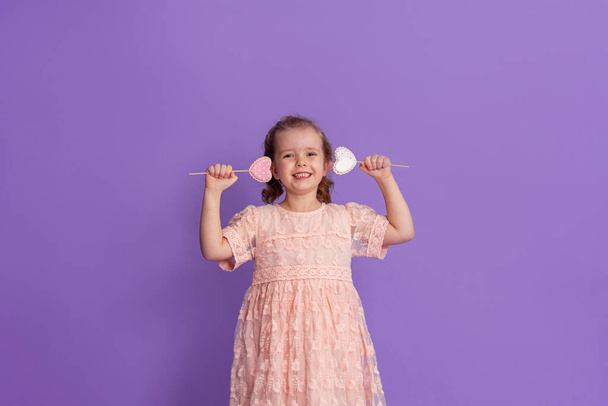 smiling little girl in a soft pink dress holds gingerbread on a stick, in the shape of hearts on a purple background in the studio. The concept of treats for children's birthday and Valentine's Day - Foto, Bild