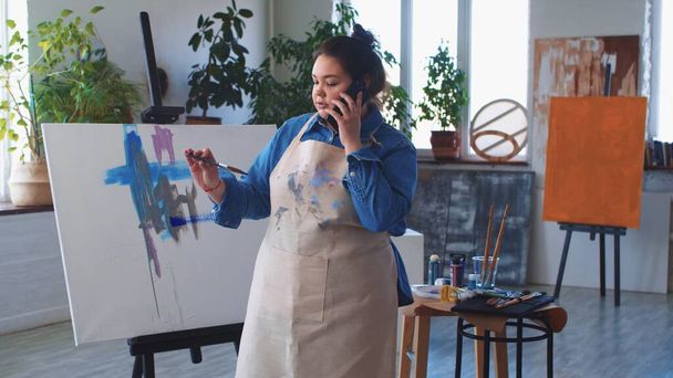 Egy nő festő művész beszél a telefonjában, miközben rajzol egy festményt. - Fotó, kép