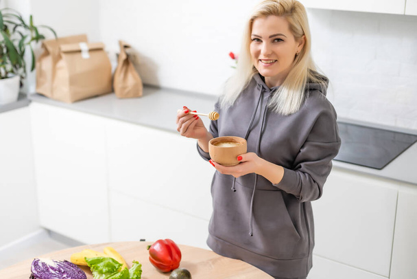 Nuori onnellinen nainen pitelee hunajaa nauttien samalla ruoanlaitto terveellistä ruokaa keittiössä. Detox ruokavalio, laihtuminen ravitsemus käsite - Valokuva, kuva