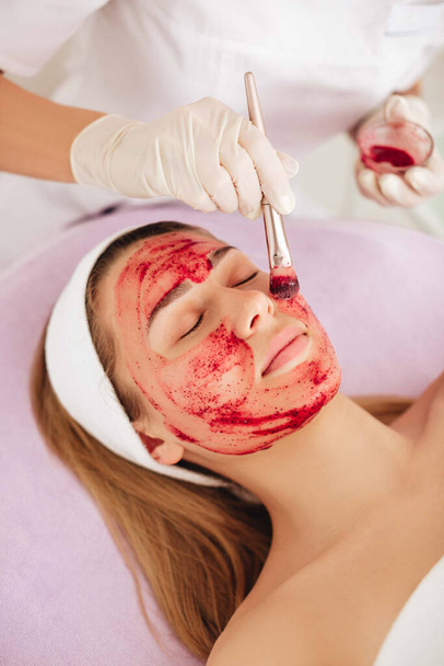 A beautiful young woman with a strawberry mask on her face lies on a couch in a beauty salon. The beautician doctor applies a facial mask to a woman's face. Professional skin care. - Photo, Image