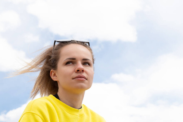 Chica joven reflexiva y auténtica con sudadera amarilla mirando a la distancia sentada afuera bajo el cielo azul y las nubes blancas. Soñar con un futuro positivo y disfrutar del concepto de vida. - Foto, imagen