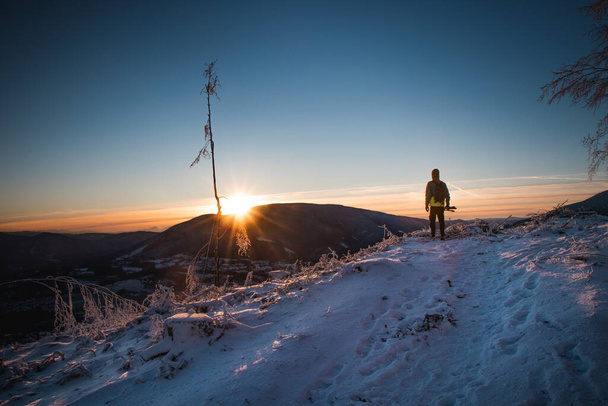 Zonsopgang in de winter in de Beskydy bergen in Oost-Bohemen. Een tiener in een kleurrijk jasje staat op de rand van een klif en de zon komt links op. Uiterste datum voor succes. - Foto, afbeelding