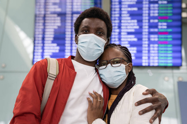 Portret van gelukkige zwarte paar reizigers in medische maskers over luchthaven terminal board achtergrond - Foto, afbeelding