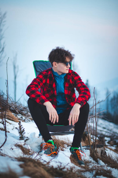 Handsome teenager aged 15-20 in a plaid red-black shirt and sunglasses on a chair by the tent. Watching sunsets in winter. Fashion photo. Candid portrait adolescent of European descent. - Photo, Image