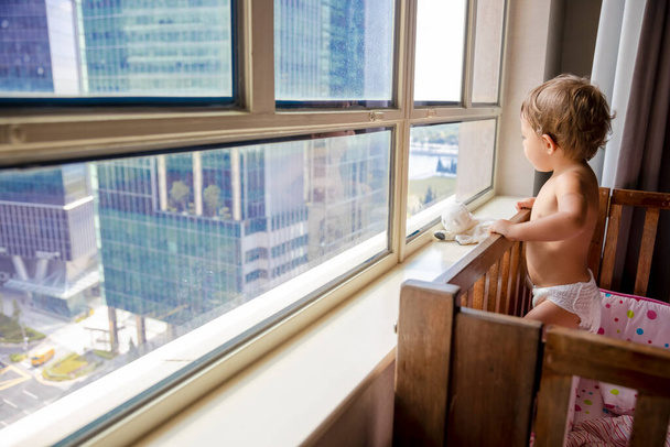 uroczy maluch patrzy przez okno na metropolię stojąc w łóżeczku. Dziecko w łóżeczku wygląda przez okno na miasto. widok z tyłu miękka ostrość - Zdjęcie, obraz