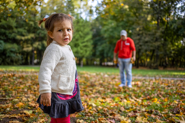 δυσαρεστημένο παιδί στο πάρκο με τον μπαμπά. Το θυμωμένο κορίτσι στο πάρκο κοιτάζει την κάμερα. Ο πατέρας και η κόρη περπατούν στο πάρκο του φθινοπώρου - Φωτογραφία, εικόνα