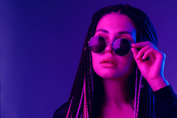Stylish young woman with braids wearing sunglasses against purple background - Zdjęcie, obraz