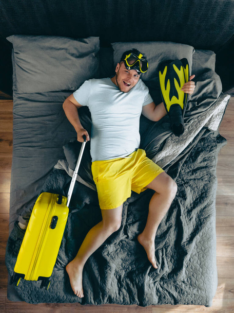 homem na cama com mala amarela segurando nadadeiras e máscara de mergulho vista superior - Foto, Imagem