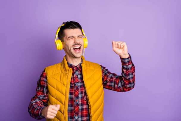 Photo of sweet funny young guy dressed yellow vest earphones singing closed eyes isolated purple color background - Foto, Bild