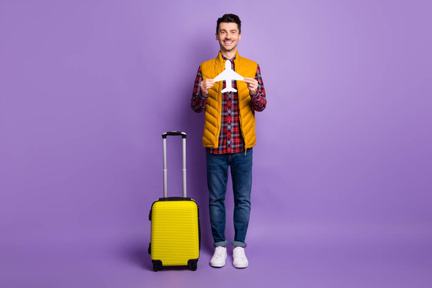 Full length photo of cute young man wear yellow sleeveless jacket holding white paper plane suitcase isolated violet color background - Zdjęcie, obraz
