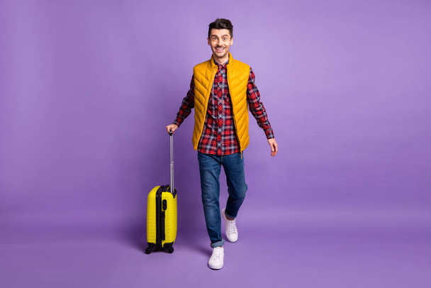 Plná délka fotografie roztomilé sladké mladý chlap oblečený žlutá vesta chůze držení zavazadel izolované fialové barvy pozadí - Fotografie, Obrázek