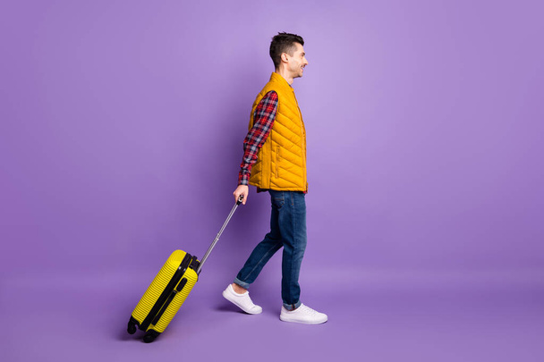 Full length photo of cheerful young man wear yellow sleeveless jacket holding suitcase walking isolated violet color background - Fotó, kép