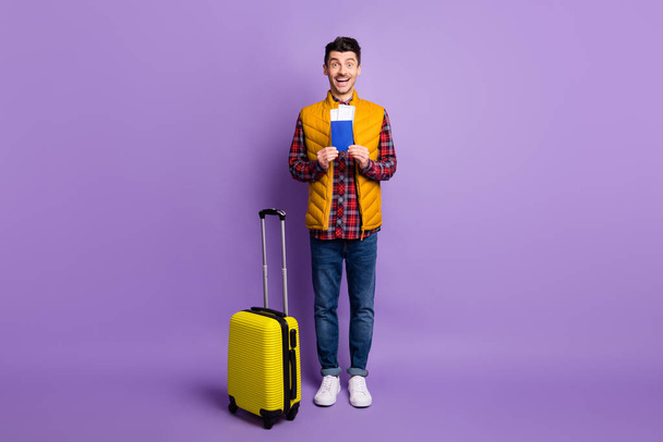 Full length photo of sweet excited young guy dressed yellow vest holding tickets luggage isolated purple color background - Φωτογραφία, εικόνα