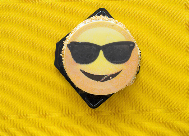 divertente torta rotonda al limone con emoji sorridente con occhiali da sole decorazione su uno sfondo strutturato giallo brillante  - Foto, immagini