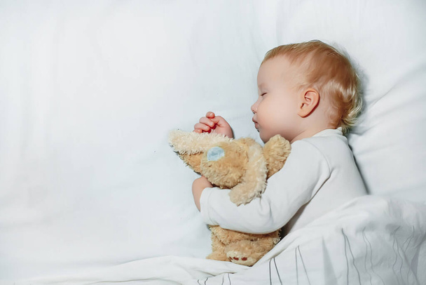 甘い小さな女の子はベッドで白いリネンで眠る。1歳の子供はおもちゃを抱きかかえて眠る。子供の概念の幸せな子供時代と睡眠の問題 - 写真・画像