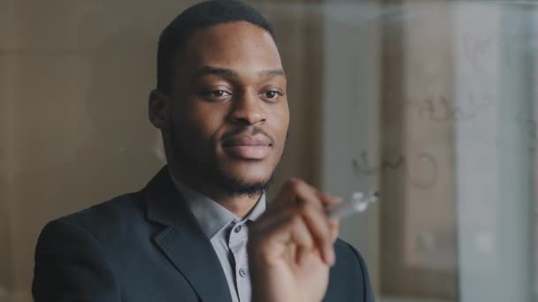 Porträt eines fokussierten afroamerikanischen Geschäftsmannes in den 30er Jahren: Manager schreibt Formel-Finanzberechnungen auf transparenter Glasplatte, analysiert Start-up, macht Innovation, erzielt Erfolg - Filmmaterial, Video