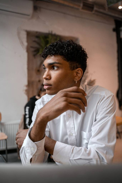 A young black man is sitting at the table with a laptop, drinking tea and using a smartphone. The guy in the cafe. Work outside the office, remote work or study concept. - Photo, Image