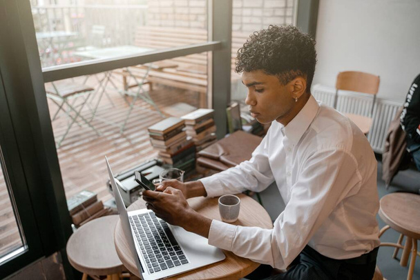 Ένας νεαρός μαύρος κάθεται στο τραπέζι με ένα φορητό υπολογιστή, πίνοντας τσάι και χρησιμοποιώντας ένα smartphone. Ο τύπος στο καφέ. Εργασία εκτός γραφείου, εξ αποστάσεως εργασία ή μελέτη έννοια. - Φωτογραφία, εικόνα
