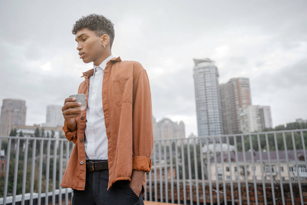 Молодой чернокожий мужчина в коричневой куртке держит в руках напиток в чашке на фоне городского горизонта или центра города. Парень пьет кофе или чай на фоне города. - Фото, изображение