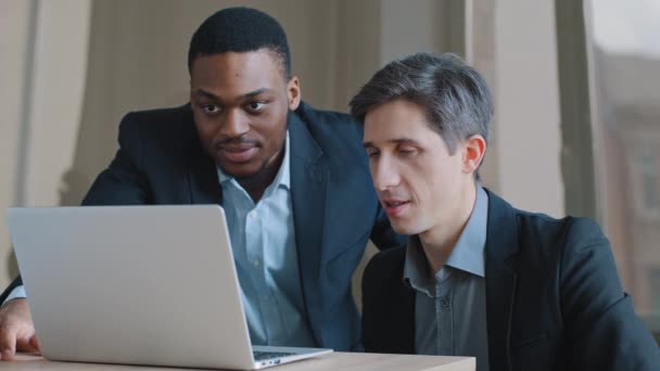 Dva zamyšlení kolegové v kanceláři hovořili o problému s online serverem, který se dívá na obrazovku notebooku pomocí počítačové aplikace, bělošský manažer zobrazující data afroamerickému obchodníkovi v síti - Záběry, video