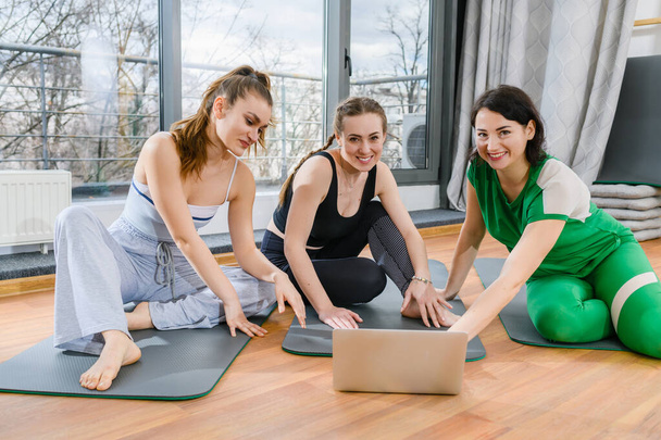 Три спортивные девушки сидят на ковриках в фитнес-студии во время тренировочных занятий по йоге онлайн - Фото, изображение