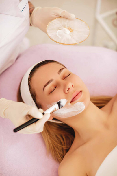 Beautician applies a white skin mask on client's face with help of brush. Beautiful woman lying and relaxing in beauty salon. Beauty anti wrinkle procedures making face moisturize.  - Photo, Image