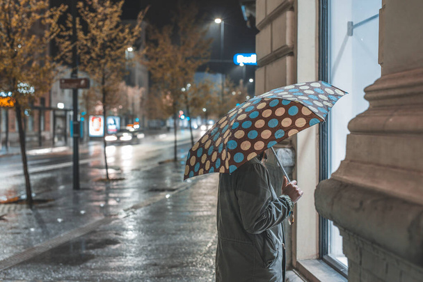 Yksinäinen nainen ikkunaostoksilla yksin yöllä sateella. Sateinen kävely läpi kaupungin. - Valokuva, kuva