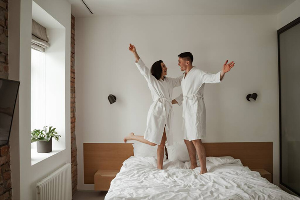 Καλημέρα ευτυχισμένο ζευγάρι με λευκά μπουρνούζια φιλιούνται και χορεύουν στο κρεβάτι στο σπίτι - Φωτογραφία, εικόνα