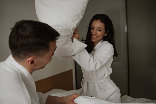 Morning happy young couple in white bathrobes are beating pillows in bed at home - Fotó, kép