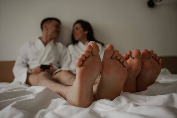 Close up of legs happy young couple in white bathrobes in bed at home - Фото, изображение
