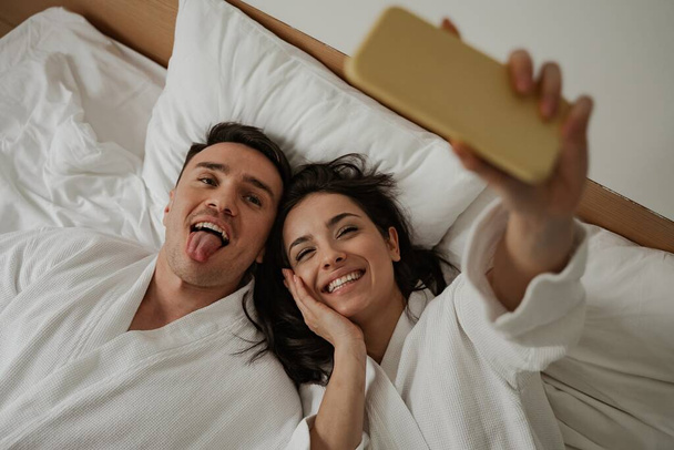 Matin de jeune couple heureux en peignoirs blancs dans le lit à la maison, faire selfie - Photo, image