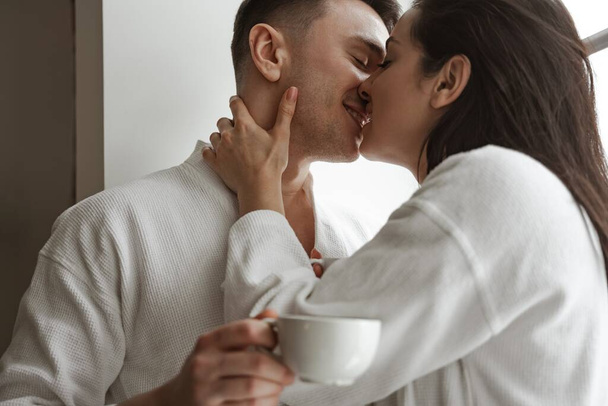 Πρωινό ευτυχισμένου ζευγαριού που φιλιέται με λευκά μπουρνούζια στο σπίτι, με ένα φλιτζάνι καφέ - Φωτογραφία, εικόνα