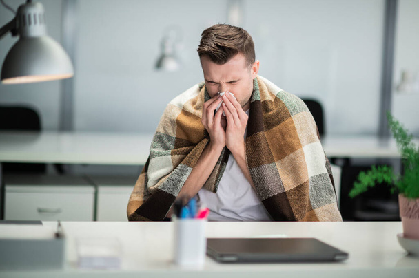 Junger Mann wird bei der Arbeit krank und pustet sich am Schreibtisch mit Serviette auf die Nase - Foto, Bild