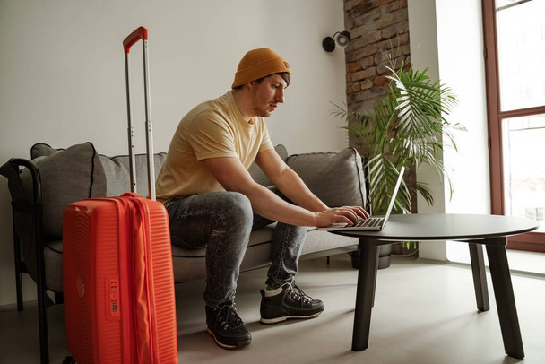 Genç bir gezgin yaz tatilinde otel odasında dizüstü bilgisayar kullanarak kanepede oturuyor. Havalı erkek, günlük giysiler içinde, bavullu. - Fotoğraf, Görsel