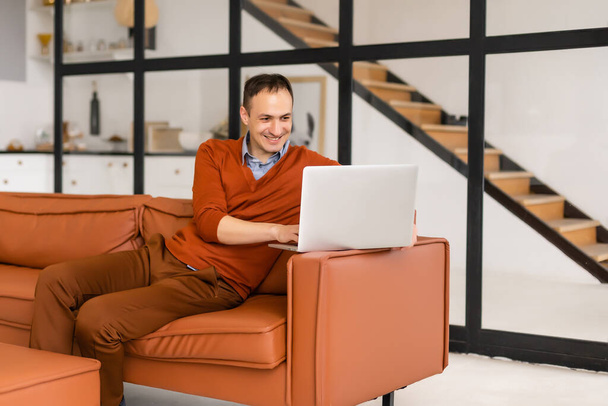 Νεαρός άνδρας χαμογελά, διαβάζει την οθόνη ενός φορητού υπολογιστή ενώ χαλαρώνει σε έναν άνετο καναπέ στο σπίτι - Φωτογραφία, εικόνα