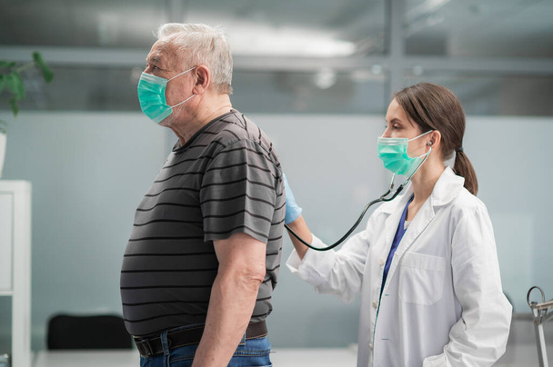 Während der ärztlichen Untersuchung führt eine Ärztin die Auskultation der Lunge eines älteren Mannes von hinten durch - Foto, Bild