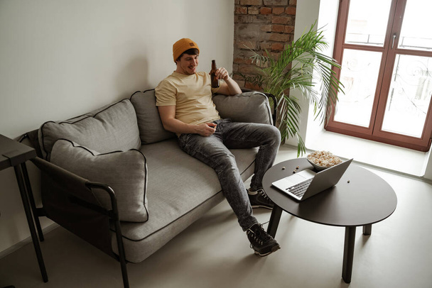 Junger Mann mit gelbem Hut trinkt Bier mit Popcorn und schaut sich im Hotelzimmer einen Film auf dem Laptop an. Männchen im Urlaub - Foto, Bild